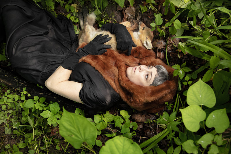 The artist wears a black dress, black elbow gloves and a fox fur shawl around her head. She is lying in a forest of bright green leaves and holds a dead baby fox in her arms.